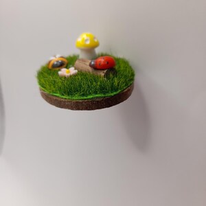 May include: A miniature fairy garden scene with a circular base covered in artificial grass. A yellow and white mushroom, a red ladybird, a bumblebee, and a small flower are arranged on the grass, creating a whimsical display.