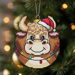 Pode incluir: Um enfeite de Natal em ponto de cruz com uma vaca Highland castanha a usar um gorro de Pai Natal vermelho e branco. A vaca tem um focinho claro, bochechas rosadas e está envolvida por um fio de luzes de Natal coloridas com um gancho dourado.