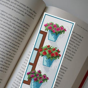 以下が含まれることがあります： ピンクと赤の花が入った青い植木鉢が3つ描かれた、手作りのクロスステッチのブックマーク。 ブックマークは茶色の梯子模様と青い縁取りがあり、開いた本の上に置かれています。