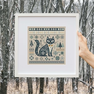Puede incluir: Arte de punto de cruz enmarcado que presenta un gato negro estilizado con pajarita, rodeado de copos de nieve, árboles y patrones geométricos en tonos azules, verdes y dorados sobre un fondo beige. La obra de arte se muestra en un marco blanco.