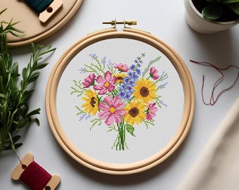 May include: A cross-stitch embroidery of a vibrant floral bouquet, featuring sunflowers, pink daisies, and blue wildflowers, displayed in a wooden embroidery hoop. Sewing supplies, including thread spools and greenery, surround the craft project.