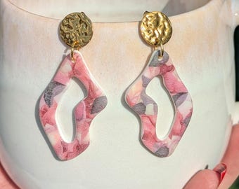 May include: A pair of pink and white patterned earrings with gold accents. The earrings are shaped like an irregular oval with a cutout in the center.