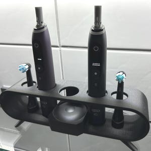 Könnte beinhalten: Schwarzer Zahnbürstenhalter mit zwei elektrischen Zahnbürsten und zwei Bürstenköpfen. Die Zahnbürsten sind schwarz und lila. Die Bürstenköpfe haben blaue und weiße Borsten. Der Halter hat eine Ladestation.