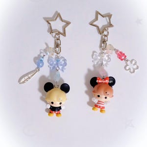 May include: Two keychains with cartoon character figures. Each keychain has a star-shaped clasp, a small bow, and beaded accents in blue and pink. One figure has blonde hair and the other has red hair. The figures are wearing black and red outfits.