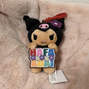 May include: A plush Kuromi character keychain featuring a black hood with a pink skull emblem and a purple flower. The character holds a rectangular sign with the text HAFA ADAI in colorful letters, decorated with small purple and white flowers.