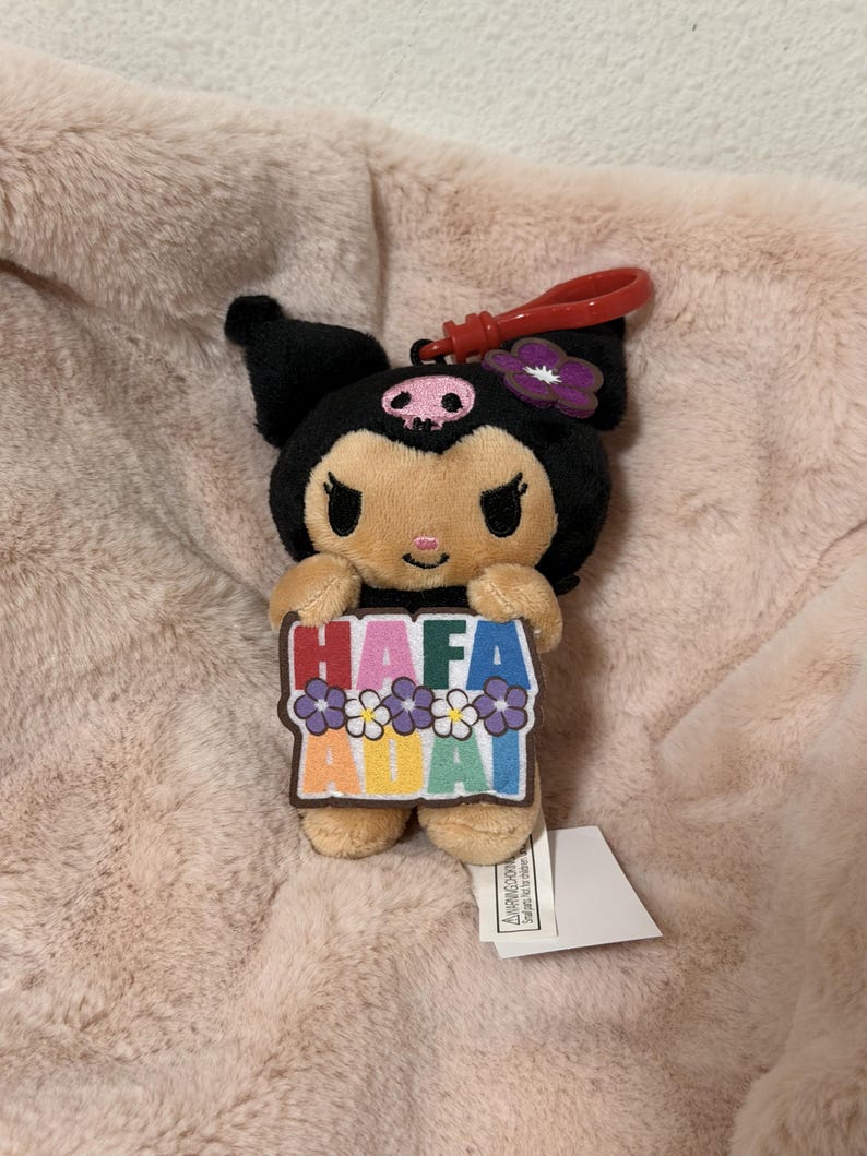 May include: A plush Kuromi character keychain featuring a black hood with a pink skull emblem and a purple flower. The character holds a rectangular sign with the text HAFA ADAI in colorful letters, decorated with small purple and white flowers.