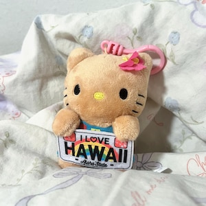 May include: A tan plush Hello Kitty keychain with a pink plastic clip and a pink flower on its head. The plush holds a white rectangular sign that reads I LOVE HAWAII with a rainbow graphic and the text Aloha State printed below it.
