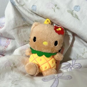 May include: A tan plush character toy featuring a yellow pineapple-patterned body, a green collar, and a small red flower accessory on its head. The character has black oval eyes and a small yellow embroidered nose, with a yellow loop on top.