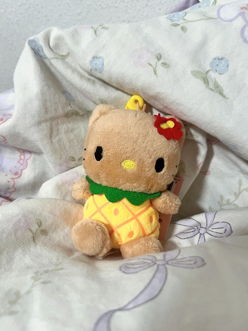 May include: A tan plush character toy featuring a yellow pineapple-patterned body, a green collar, and a small red flower accessory on its head. The character has black oval eyes and a small yellow embroidered nose, with a yellow loop on top.