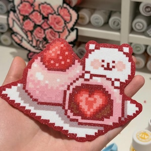 May include: A glittery pixel art sticker featuring a white bear character next to a pink mochi dessert. The mochi is topped with a red strawberry and contains a red heart-shaped filling. The design uses a vibrant red and pink color palette.