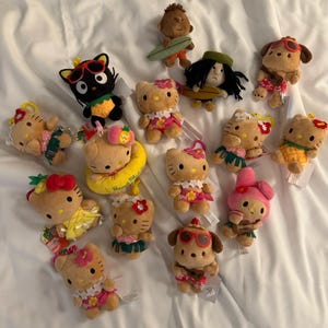 May include: A collection of plush toys featuring various characters, including Hello Kitty, a black cat, and a dog, all dressed in Hawaiian-themed outfits. Some toys hold surfboards or wear sunglasses. The toys are set against a white background.