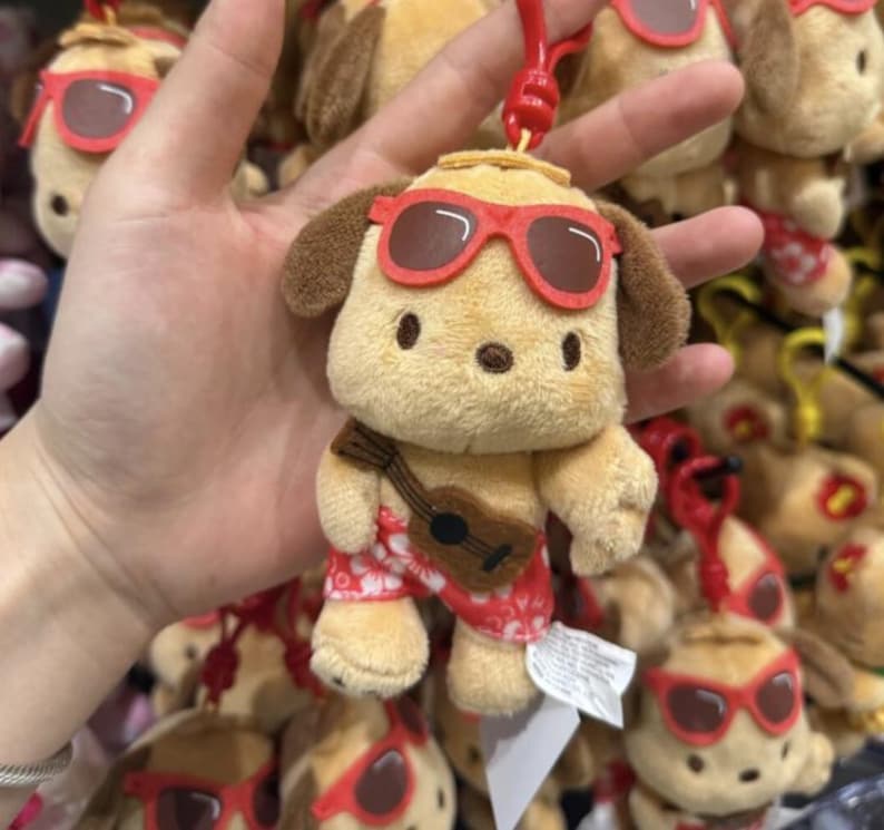 May include: A plush dog keychain featuring a tan character wearing red sunglasses and red floral patterned shorts, holding a small brown guitar. The toy has a red plastic clip attached to the top of its head and a white fabric tag on its side.