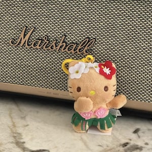 May include: A plush Hello Kitty keychain featuring a tan character wearing a white and red flower crown, a pink seashell top, and a green hula skirt. The figure has a yellow nose, brown eyes, and a yellow plastic clip attached to the top.