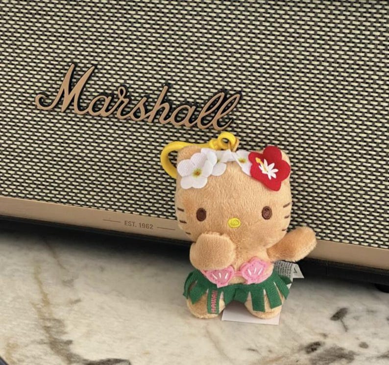 May include: A plush Hello Kitty keychain featuring a tan character wearing a white and red flower crown, a pink seashell top, and a green hula skirt. The figure has a yellow nose, brown eyes, and a yellow plastic clip attached to the top.