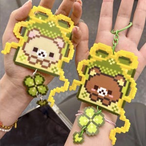 May include: Two pixel art keychains made of fused plastic beads. Each features a square bear character wearing a green hood with yellow accents, attached to a four-leaf clover charm. One bear is light beige, the other is brown, both with pixelated faces.