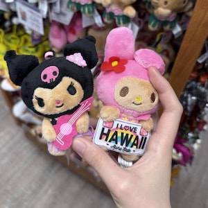 May include: Two plush toys featuring Kuromi and My Melody. Kuromi is black with a pink skull and holds a pink guitar. My Melody is pink with a red flower and holds a white sign that reads I LOVE HAWAII with a rainbow and the text Aloha State.