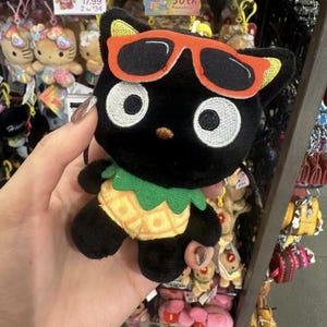 May include: A plush Chococat character keychain featuring a black cat with large white eyes, wearing orange-rimmed sunglasses and a yellow pineapple-patterned body with a green leafy collar. The toy has a small loop at the top for hanging.