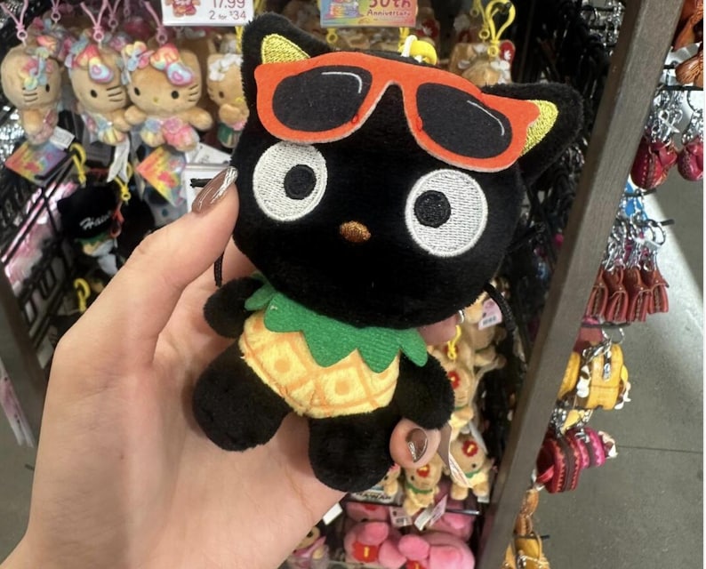 May include: A plush Chococat character keychain featuring a black cat with large white eyes, wearing orange-rimmed sunglasses and a yellow pineapple-patterned body with a green leafy collar. The toy has a small loop at the top for hanging.