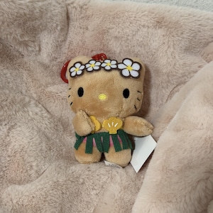 May include: A tan plush character keychain featuring a yellow nose, black eyes, and whiskers. The character wears a white flower crown, a yellow shell-shaped top, and a green grass skirt, with a red plastic clip attached to the top.