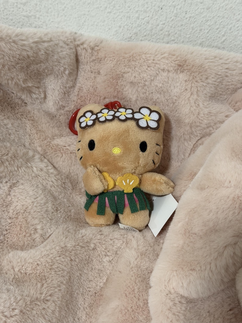 May include: A tan plush character keychain featuring a yellow nose, black eyes, and whiskers. The character wears a white flower crown, a yellow shell-shaped top, and a green grass skirt, with a red plastic clip attached to the top.