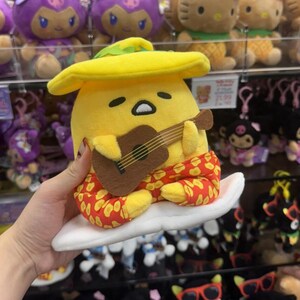 May include: A yellow Gudetama plush toy wearing a yellow hat and red patterned pants, sitting on a white surfboard while holding a brown acoustic guitar. The character has a sleepy expression with half-closed eyes and a small mouth.