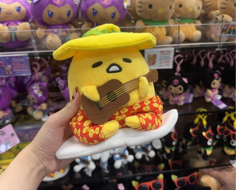 May include: A yellow Gudetama plush toy wearing a yellow hat and red patterned pants, sitting on a white surfboard while holding a brown acoustic guitar. The character has a sleepy expression with half-closed eyes and a small mouth.