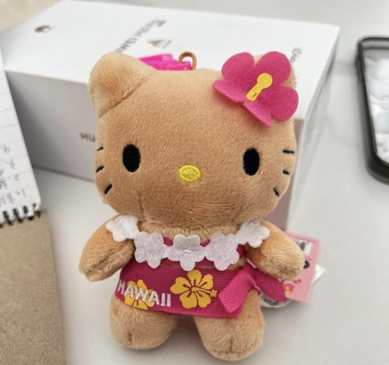 May include: A tan plush toy of a cat character with black eyes and whiskers, a yellow nose, and a pink flower accessory on its head. It wears a white floral lei and a pink dress with yellow hibiscus flowers and the text HAWAII printed on it.