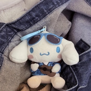 May include: A plush toy of a white character with long ears, wearing blue sunglasses and a blue floral shirt, holding a small brown ukulele. The character has pink cheeks and a blue embroidered mouth. A light blue plastic keychain is attached.