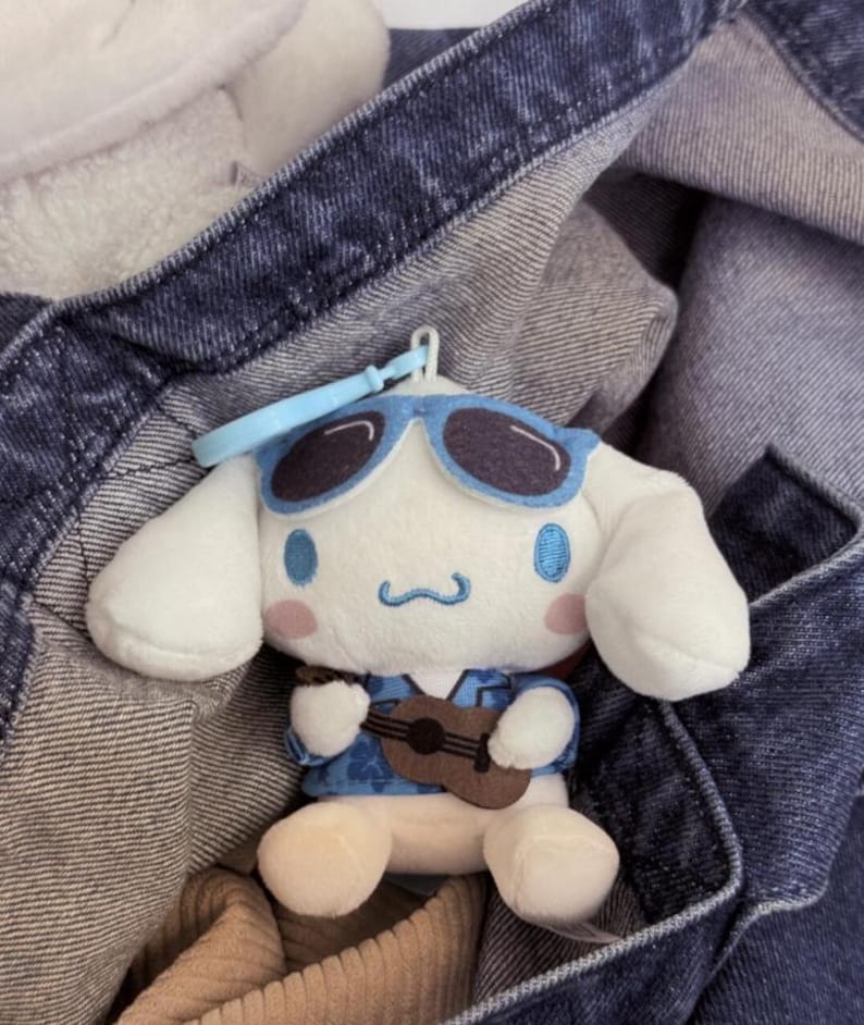 May include: A plush toy of a white character with long ears, wearing blue sunglasses and a blue floral shirt, holding a small brown ukulele. The character has pink cheeks and a blue embroidered mouth. A light blue plastic keychain is attached.