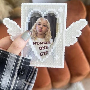 May include: A white pixel art frame made of beads featuring a heart shape and wings. The frame holds a photograph of a person with blonde hair wearing a white shirt with the text NUMBER ONE GIRL printed in dark red letters on the front.