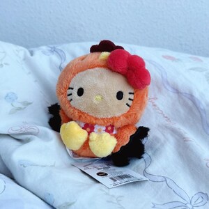 May include: A plush Hello Kitty toy dressed in an orange chicken costume with a red bow, yellow feet, and a small red comb on top. The character has black whiskers, a yellow nose, and a white bib with a red and pink floral pattern.