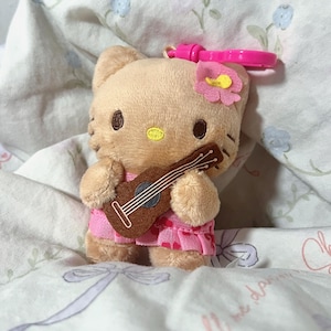 May include: A plush Hello Kitty keychain featuring a tan cat character holding a brown ukulele. The character wears a pink skirt with a heart pattern and a pink flower accessory on its head. A bright pink plastic clip is attached to the top.