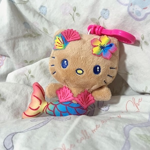 May include: A tan plush character keychain featuring a mermaid tail with blue, green, and yellow scales. The character has blue embroidered eyes, a yellow nose, and wears a pink seashell top, a pink shell hair clip, and a rainbow flower.