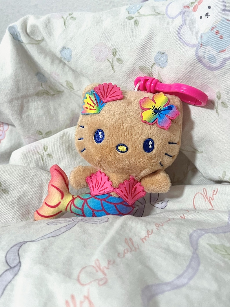 May include: A tan plush character keychain featuring a mermaid tail with blue, green, and yellow scales. The character has blue embroidered eyes, a yellow nose, and wears a pink seashell top, a pink shell hair clip, and a rainbow flower.