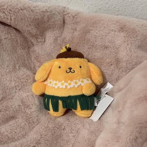 May include: A yellow plush dog character wearing a brown beret with a small yellow flower, a white daisy lei around its neck, and a dark green grass-style skirt. The plush toy has embroidered facial features and a white product tag attached.