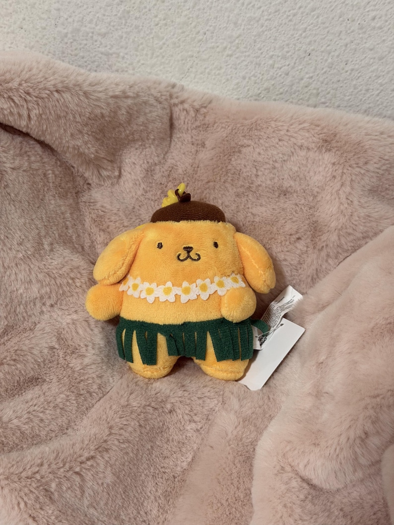 May include: A yellow plush dog character wearing a brown beret with a small yellow flower, a white daisy lei around its neck, and a dark green grass-style skirt. The plush toy has embroidered facial features and a white product tag attached.