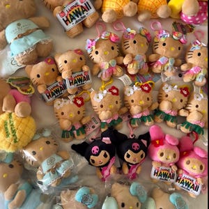 May include: A collection of Hello Kitty and Kuromi plush toys, many featuring Hawaii-themed accessories like pineapple costumes, floral leis, and surfboards. Several plushies have tags that read I LOVE HAWAII.