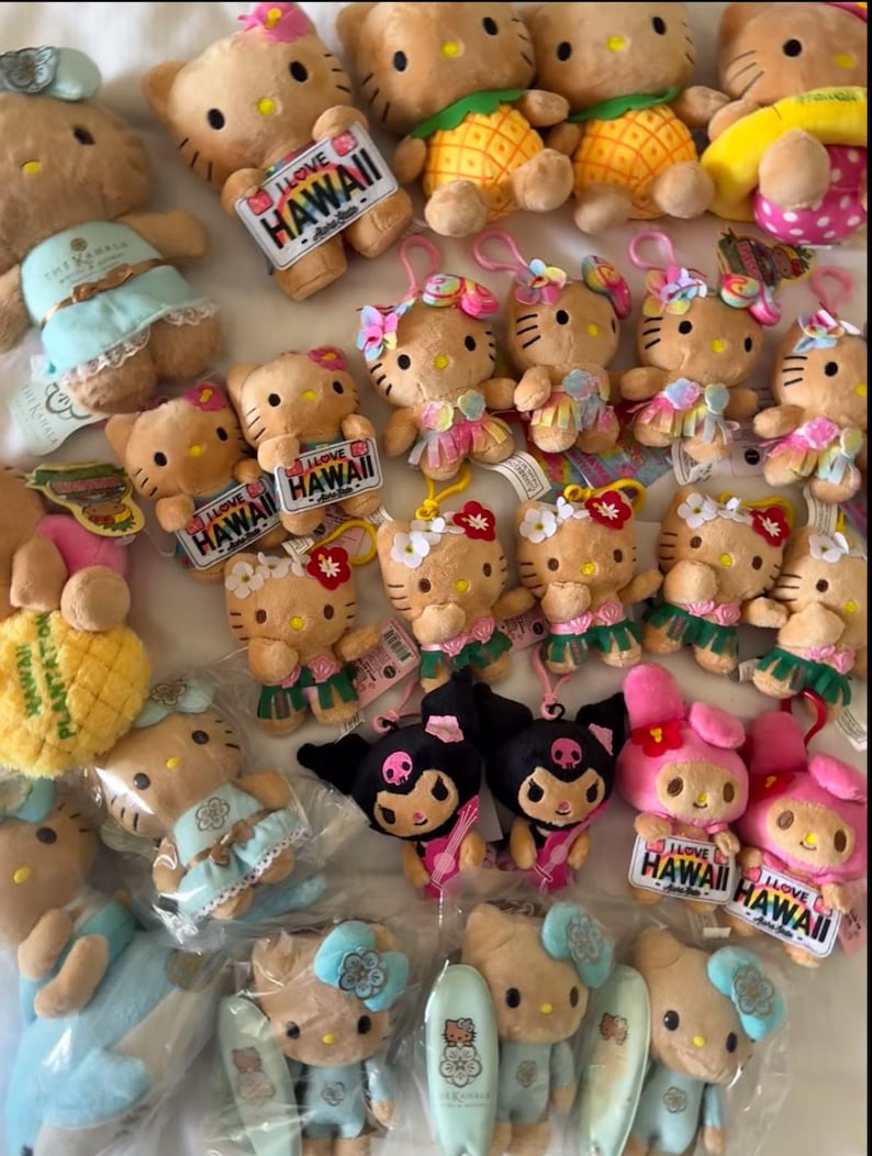 May include: A collection of Hello Kitty and Kuromi plush toys, many featuring Hawaii-themed accessories like pineapple costumes, floral leis, and surfboards. Several plushies have tags that read I LOVE HAWAII.