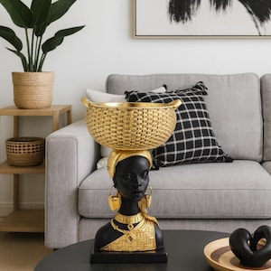 Puede incluir: Busto decorativo negro y dorado de una mujer con una cesta en la cabeza. El busto tiene joyas doradas y un tocado dorado. La cesta es dorada y tiene un diseño tejido. El busto está sobre una base negra.