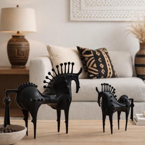 May include: Two black horse sculptures with elongated legs and vertical spikes along the mane. The sculptures are displayed on a light wood table. The background features a neutral-toned sofa and decorative items. The sculptures are made of metal.