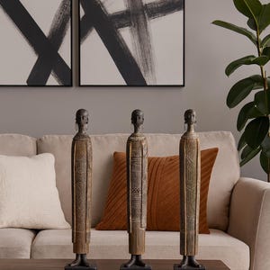 May include: Three carved wooden statues with intricate detailing, displayed on a dark wooden table. The statues, with their slender, elongated forms and dark bases, are positioned in front of a beige sofa with decorative cushions. Abstract black and white artwork adorns the wall.
