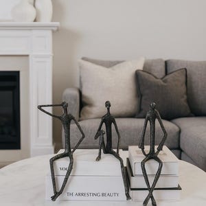 Puede incluir: Tres esculturas de metal oscuro de figuras sentadas sobre una mesa de centro de mármol blanco. Las figuras están colocadas encima de libros blancos con el texto "THE ARRESTING BEAUTY". La mesa tiene un borde de metal dorado.