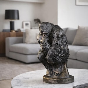 May include: A bronze-colored gorilla sculpture in a pensive pose, resting on a round, marble-topped coffee table. The sculpture has detailed fur texture and is set against a neutral-toned living room.