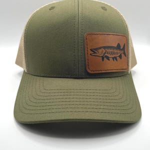 May include: Olive green trucker hat with a tan mesh back and a curved brim. A brown leather patch on the front features a black fish illustration. The hat has a structured crown and a snapback closure.