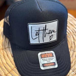 May include: Black trucker hat with a mesh back and a foam front. The hat features a white rectangular patch with the handwritten text "Let them" and a heart symbol. The hat has a durable snap closure.
