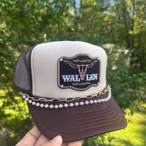 May include: A brown and tan trucker hat with a mesh back. The hat features a patch with a steer skull and the word "WAL LEN". A gold chain and pearl strand adorn the brim.