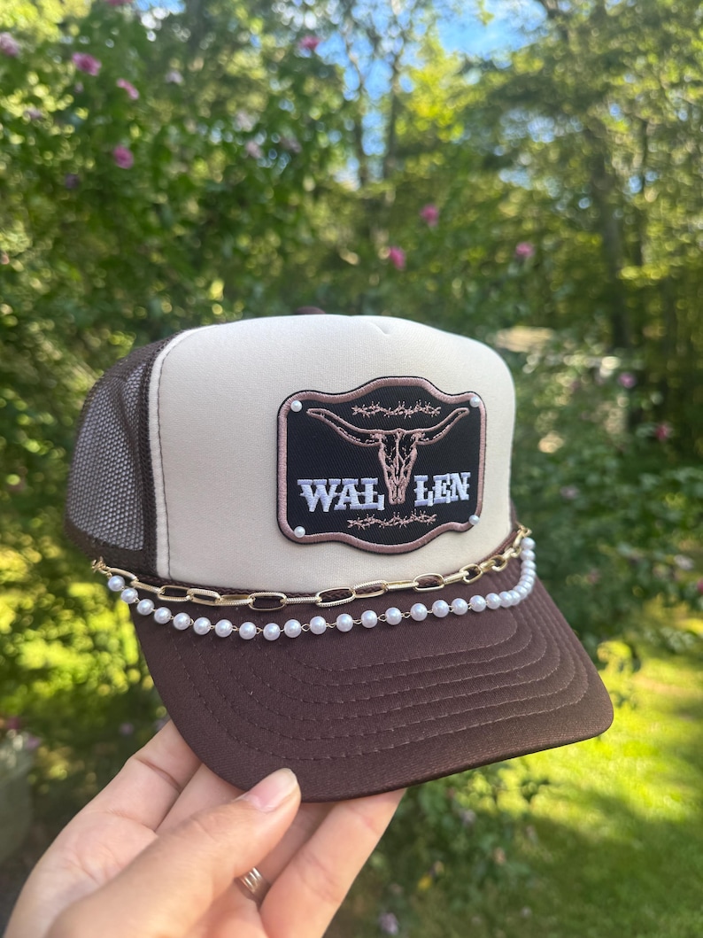 May include: A brown and tan trucker hat with a mesh back. The hat features a patch with a steer skull and the word "WAL LEN". A gold chain and pearl strand adorn the brim.