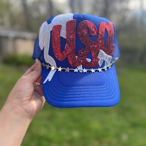 May include: A blue trucker hat with the word "USA" in red sequins. The hat features white bows and a silver star chain along the brim. The hat is a patriotic accessory.