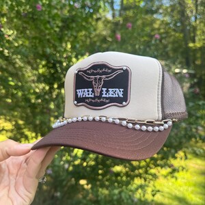 May include: A brown and tan trucker hat with a brown brim and mesh back. The hat features a black patch with a steer skull design and the word "WAL LEN" in white. A chain of pearls and gold-colored links adorns the brim.