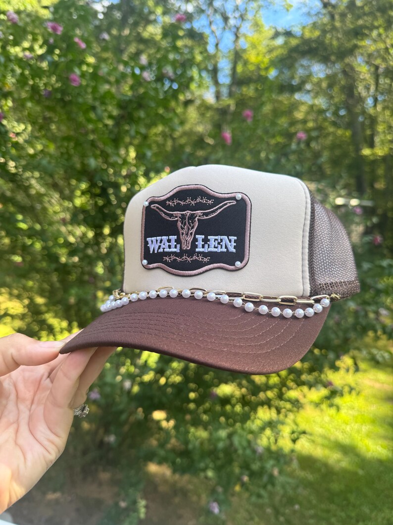 May include: A brown and tan trucker hat with a brown brim and mesh back. The hat features a black patch with a steer skull design and the word "WAL LEN" in white. A chain of pearls and gold-colored links adorns the brim.